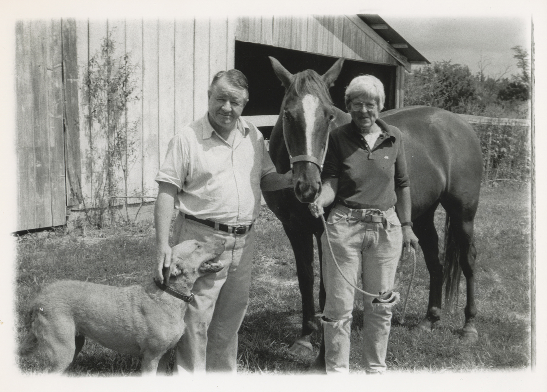 Old photo of a man and woman with a dog and horse, standing in front of a barn.
