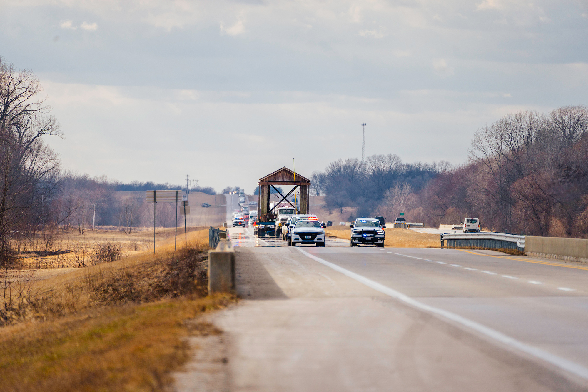 A large covered bridge on a trailer travels toward the viewer via a large oversized trucking rig, preceded by police and follow cars.