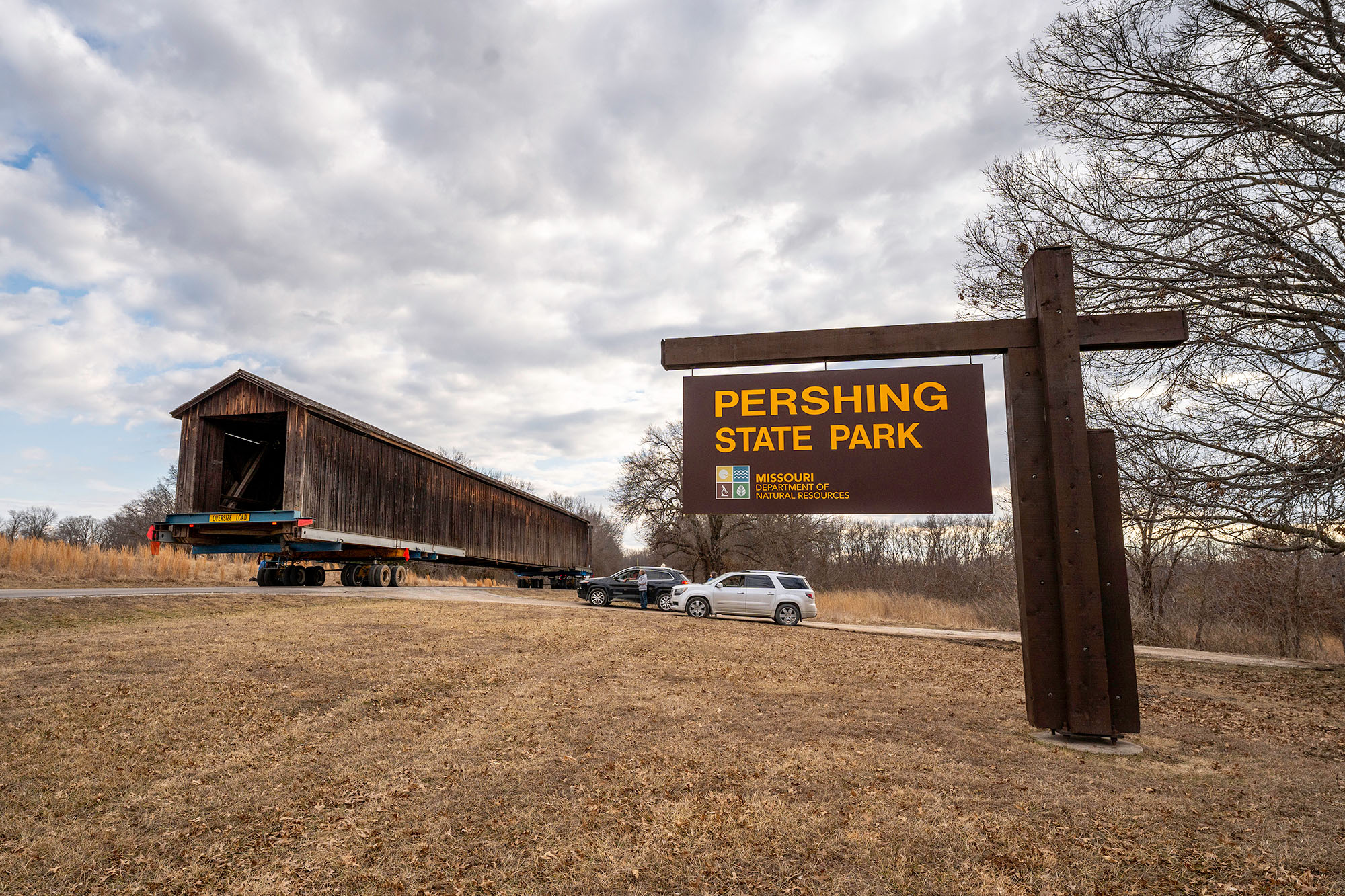 Sign for Pershing State Park in the foreground with a covered bridge on an oversized trucking rig entering the park in the background.