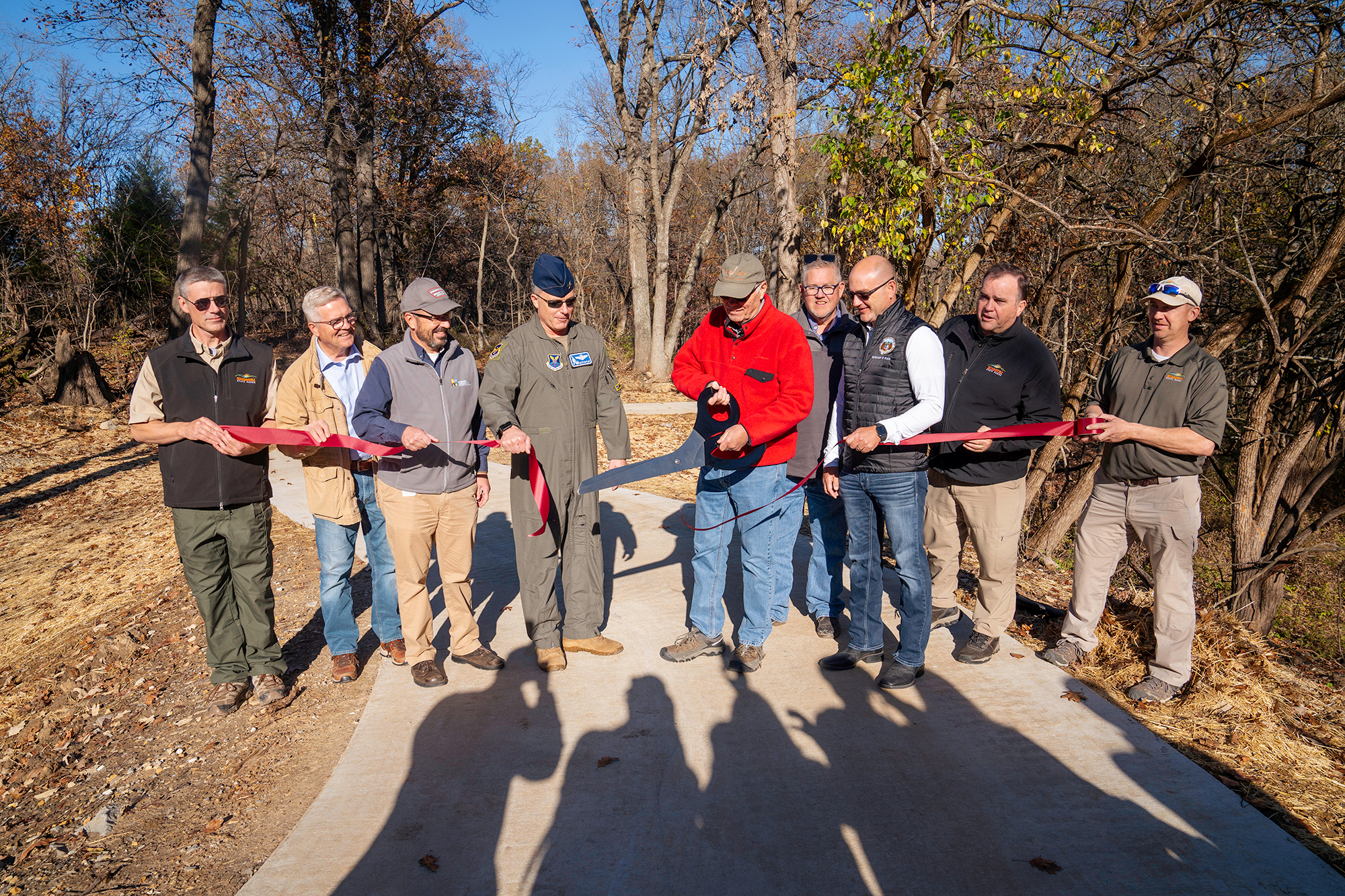 A man with giant scissors cuts through a ribbon held by a group of people on a trail.
