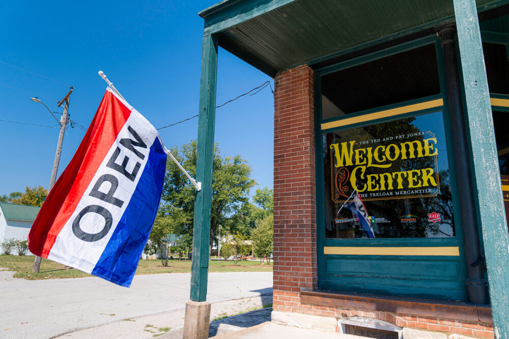 Front corner of a building with "Welcome Center" on the window. To the left of the window a Post with an "Open" flag hanging.