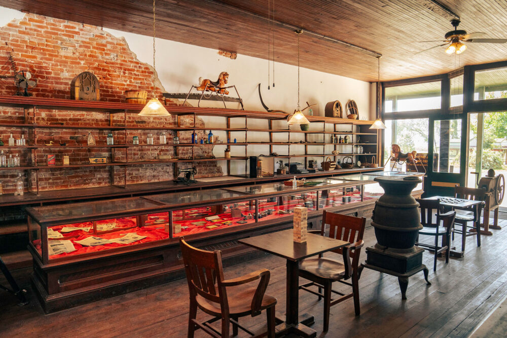 Interior of a shop, with shelves along the wall displaying vintage items. Display cases and small tables with chairs fill the remaining space.