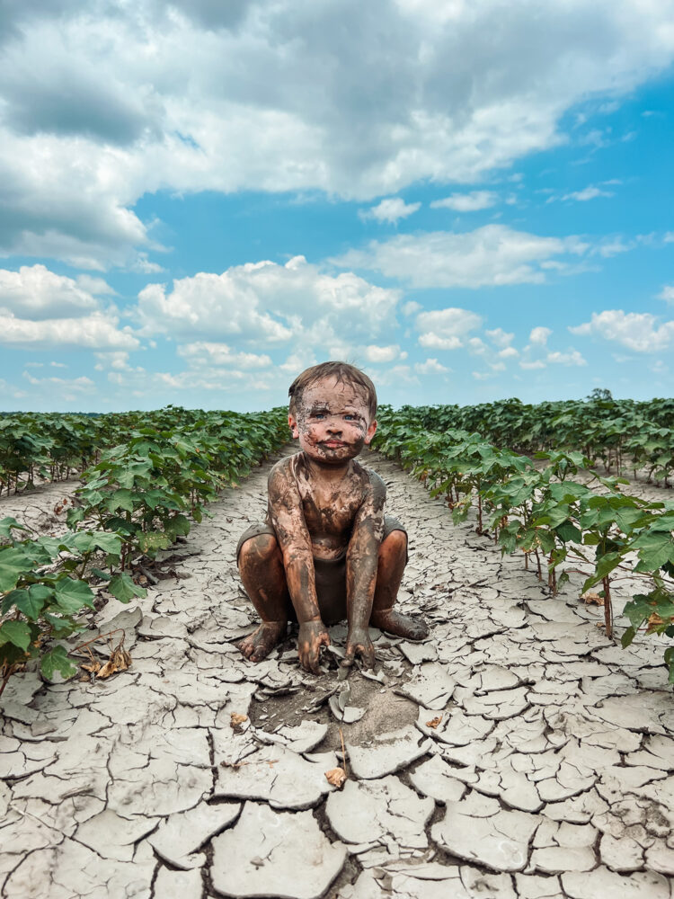 A small boy covered in mud squats between rows of cotton plants. The ground under him is dry and cracked, which makes you wonder where the mud came from.