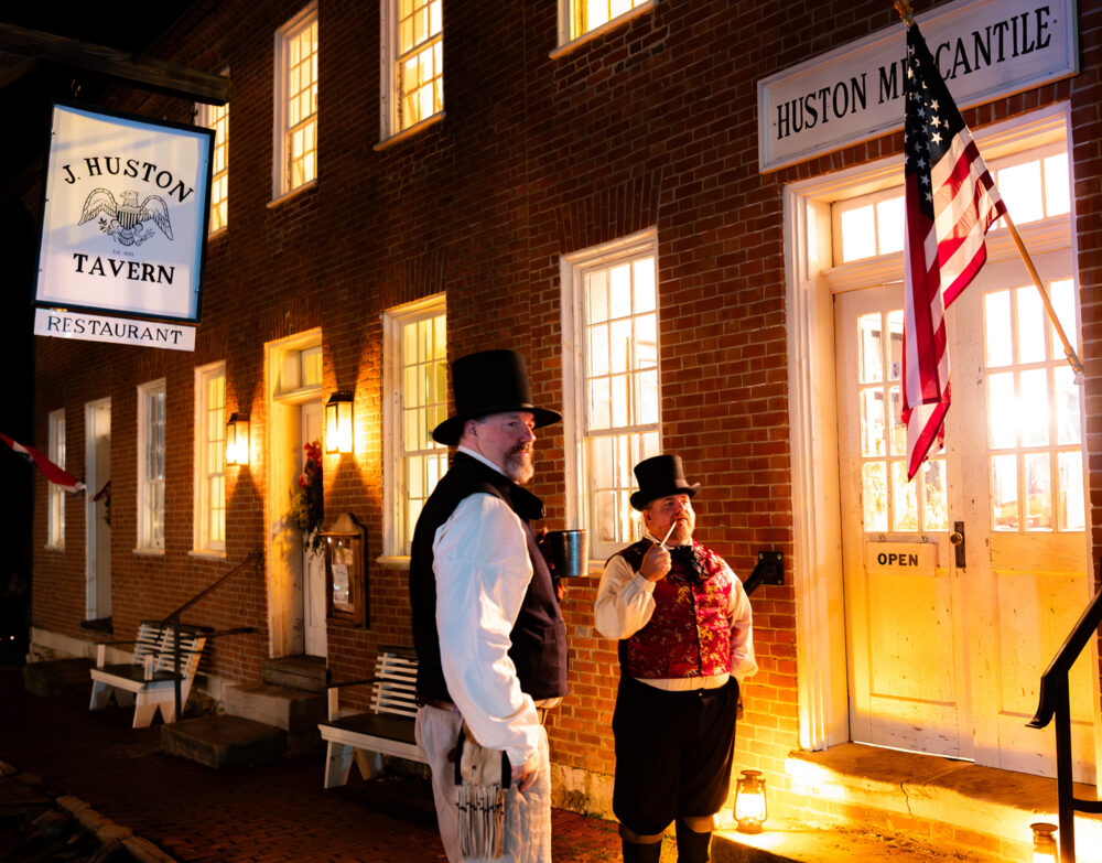 Two men dressed in colonian dress, including top hats, stand in front of a brick building. A Sign above them reads, "J. Huston Tavern". One of the men is smoking a clay pipe and the other is holding a tin mug.