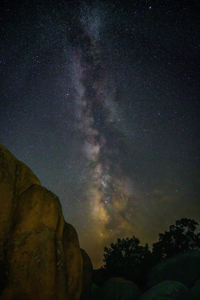 A strip of cloudy light trails upward in a sky filled with stars. In the bottom left of the frame is the edge of a large granite boulder.