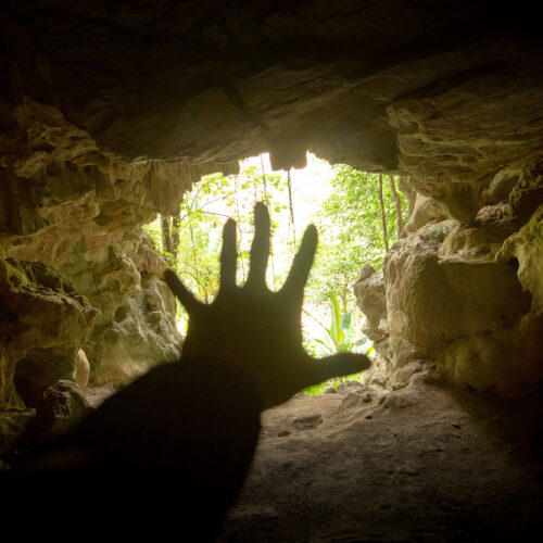 The interior of a cave facing the bright opening, obscured by the silhouette of a hand held up fingers splayed.