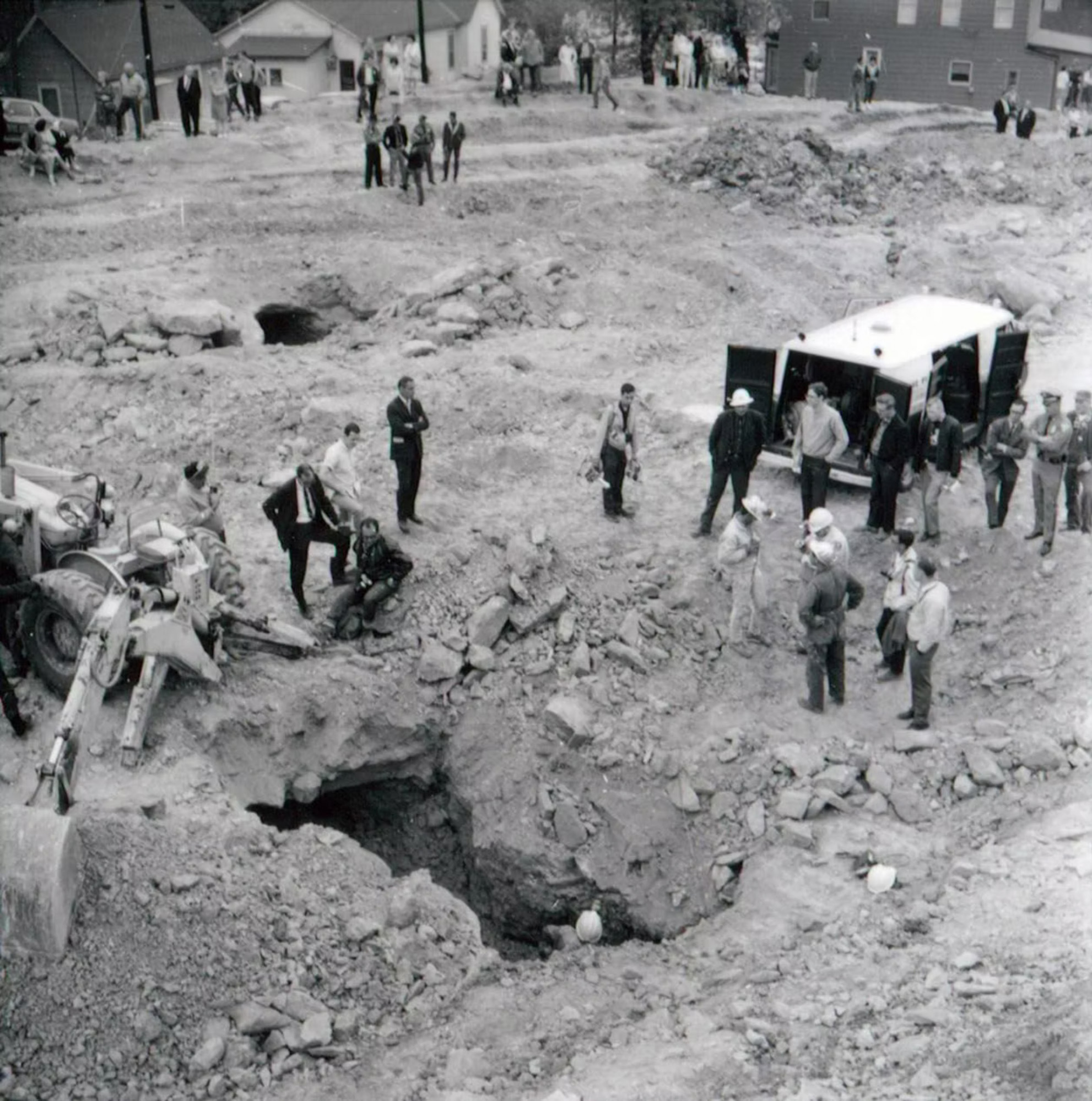 Photo from 1967, showing a large opening in the ground surrounded by people and construction equipment, one person stands inside only their head and shoulder in view.
