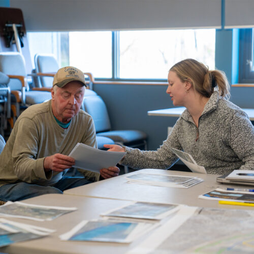 Two people sit at a table with various papers and maps, discussing a piece of paper being held by one of them.