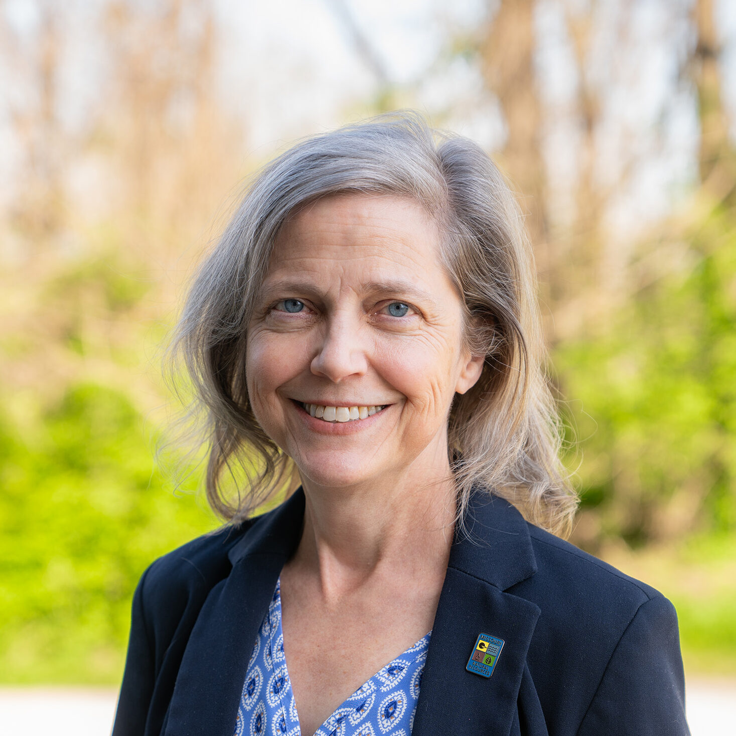 A person smiling at the camera in front of a wooded area.