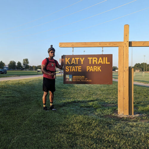 A person stands in front of a state park sign in an open area, the sign reads "Katy Trail, State Park, Missouri Department of Natural Resources."