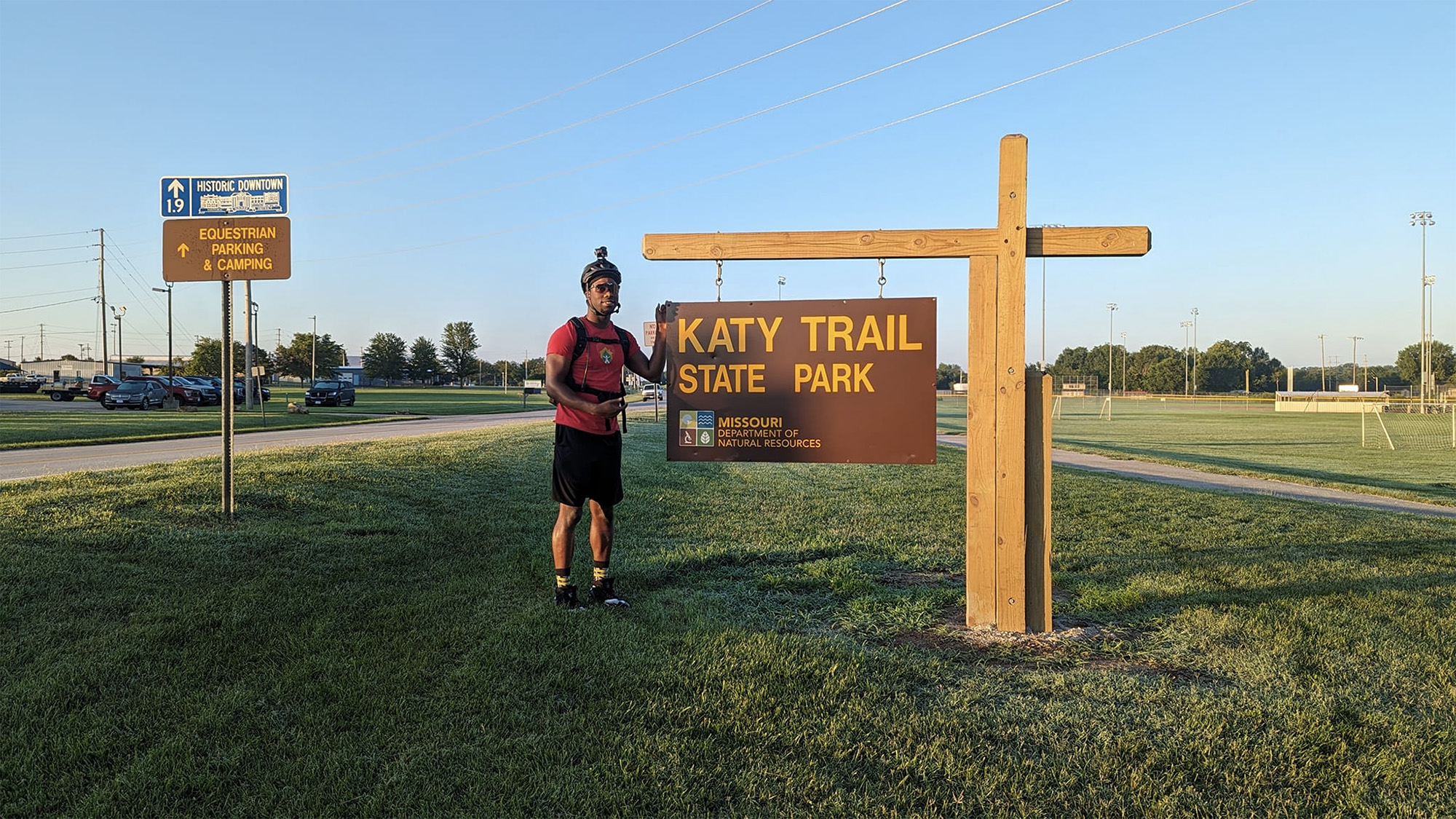 A person stands in front of a state park sign in an open area, the sign reads "Katy Trail, State Park, Missouri Department of Natural Resources."