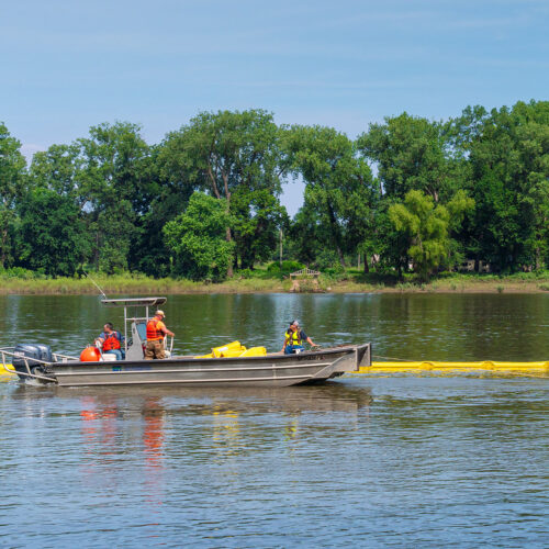 People in a flat open boat with low sides float next to a line of floating containment bouys on a large lake next to a wooded area.