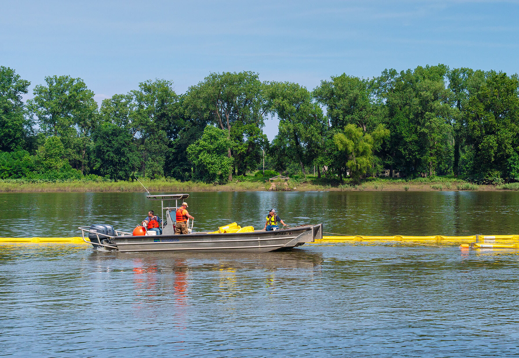 People in a flat open boat with low sides float next to a line of floating containment bouys on a large lake next to a wooded area.