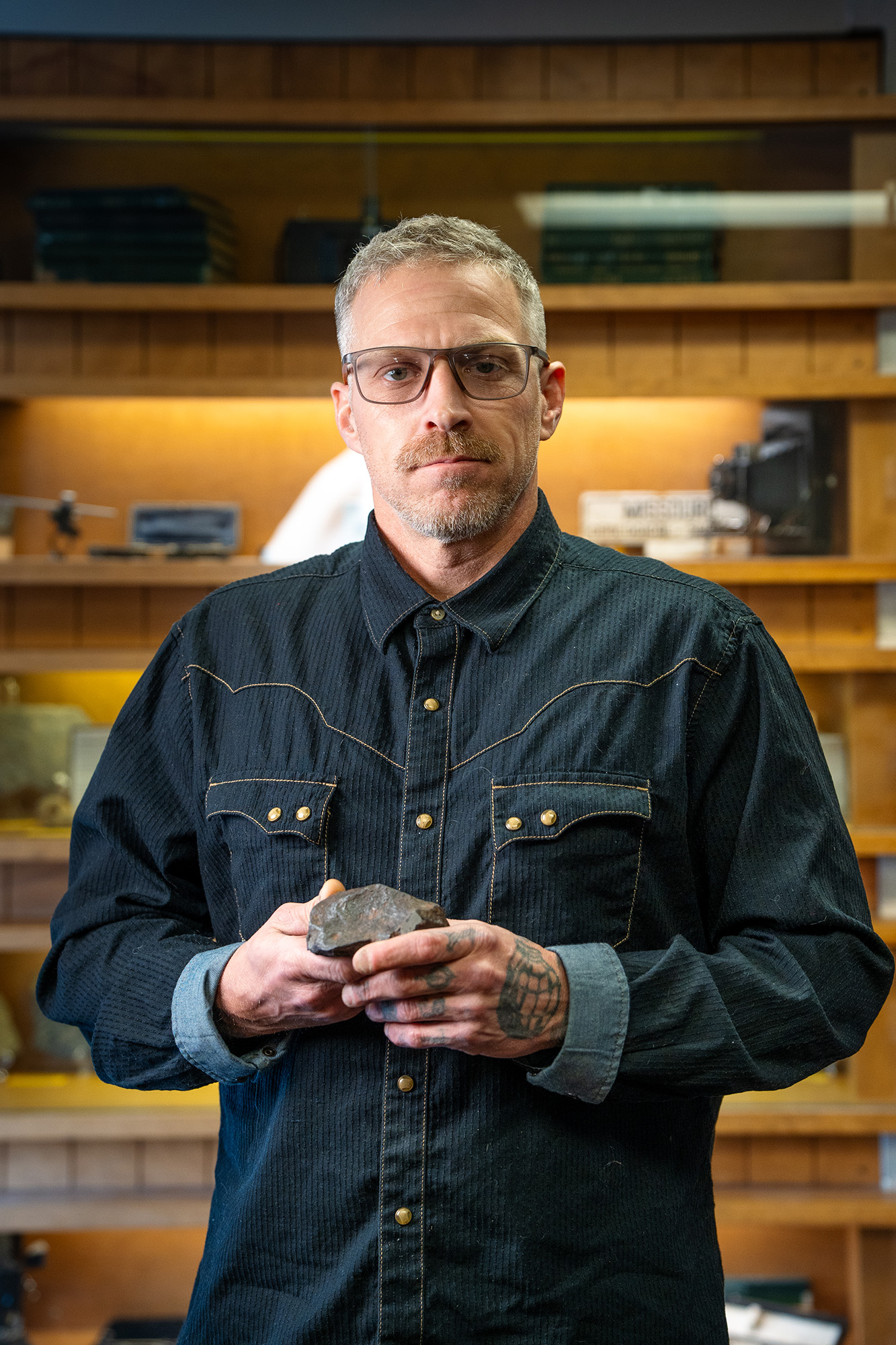 A person smiles at the camera holding a meteorite, standing in front of wooden display cases.