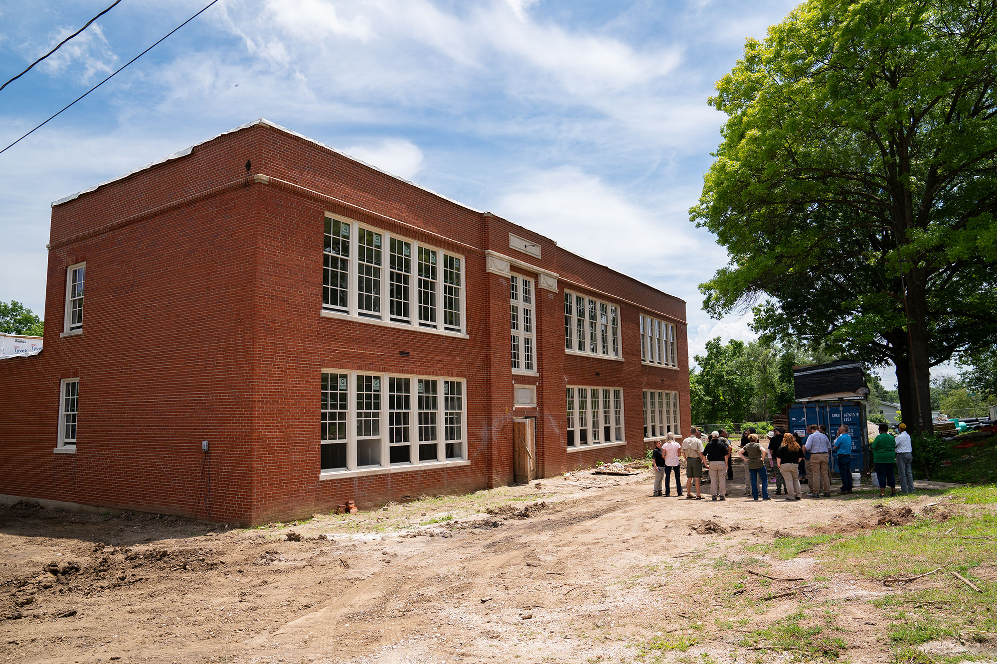 A large 2 story brick building, surrounded by freshly packed dirt stands next to a group of about 20 people gathered under a tree listening to someone speak.