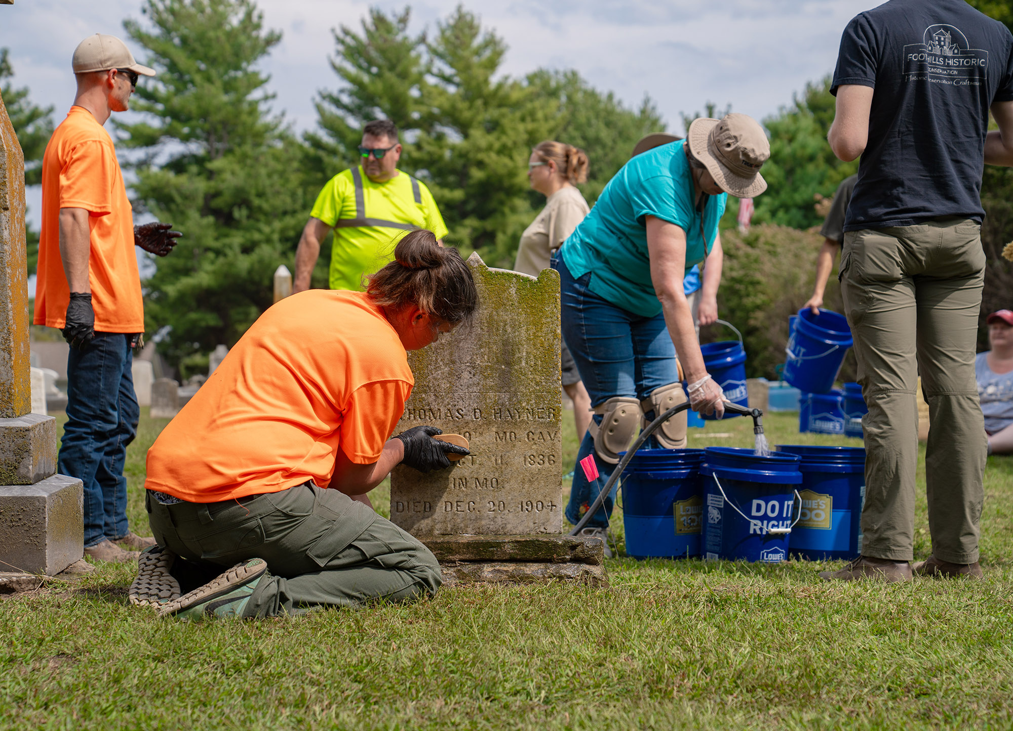 People with brushes and buckets are scattered around a cemetery in varying positions cleaning gravestones and spraying water.