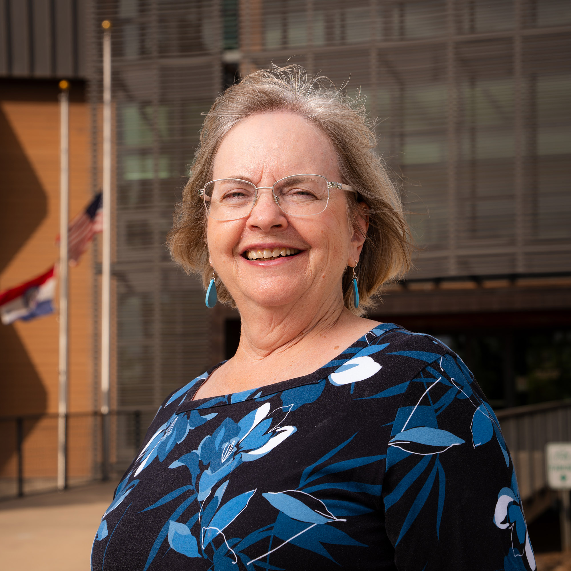 A woman stands in front of an office building. The U.S. and Missouri flags fly in the background.