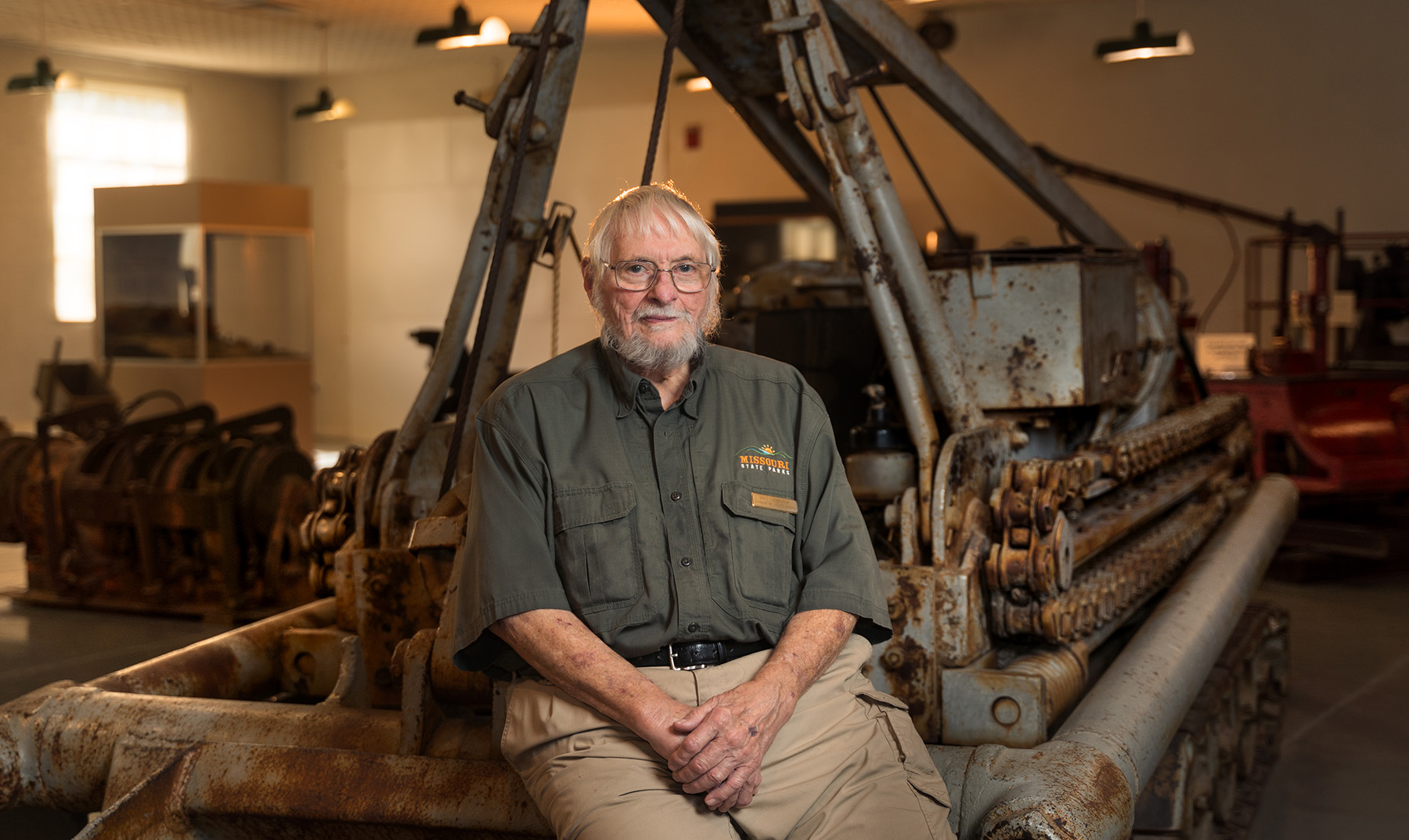 A man in a Missouri State Parks uniform sits in a room filled with old mining equipment. The equipment is made of heavy steel, with peeling paint and rusty sections.