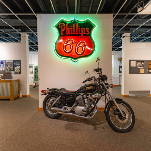 A classic motorcycle is displayed below a Phillips 66 neon sign, surrounded by other museum displays.