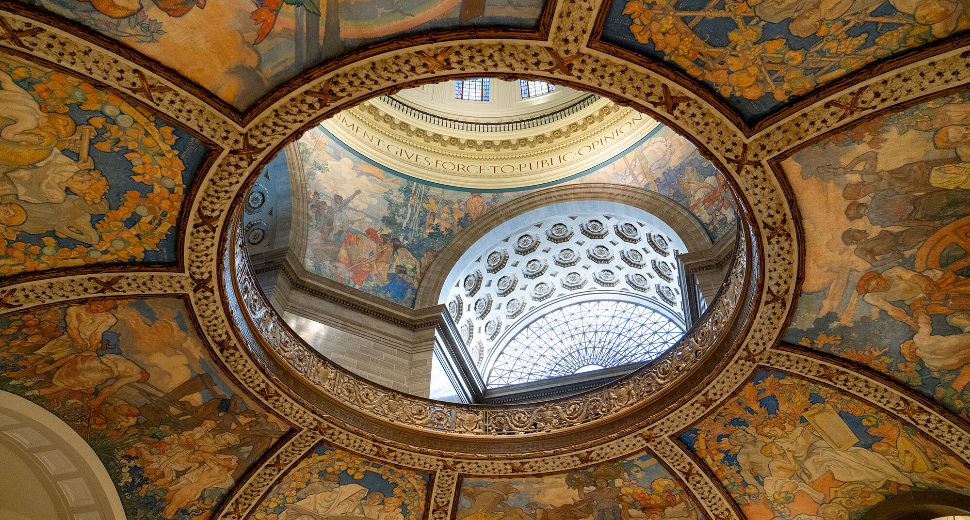 The interior of a two-tiered dome viewed from below. Fresco paintings embellish the lower tier. The upper tier features carved stone walls with geometric patterns, accented by more fresco paintings.