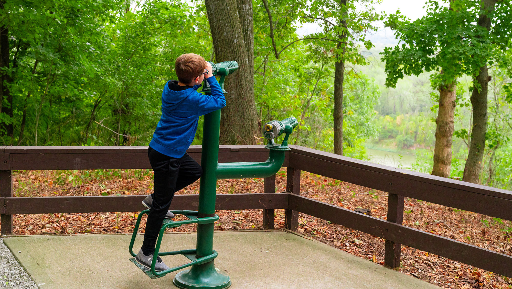 A young child looks into the distance through a monocular scope. The child is surrounded by bright open woods.