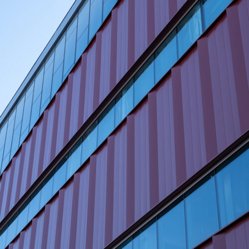 The exterior of a new building with large rows of windows and metal siding viewed from the ground set against a clear sky.