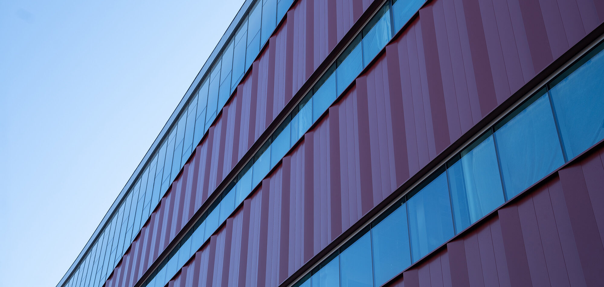 The exterior of a new building with large rows of windows and metal siding viewed from the ground set against a clear sky.