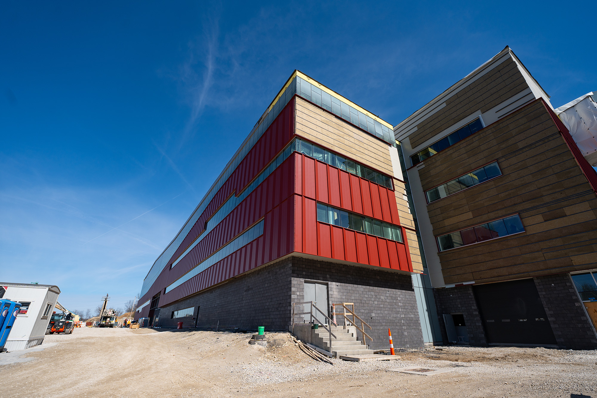 Exterior of a large building, strips of glass windows, and metal siding, sit atop a brick stone foundation. Finishing touches have not been made as construction equipment and cones sit where landscaping will later be.