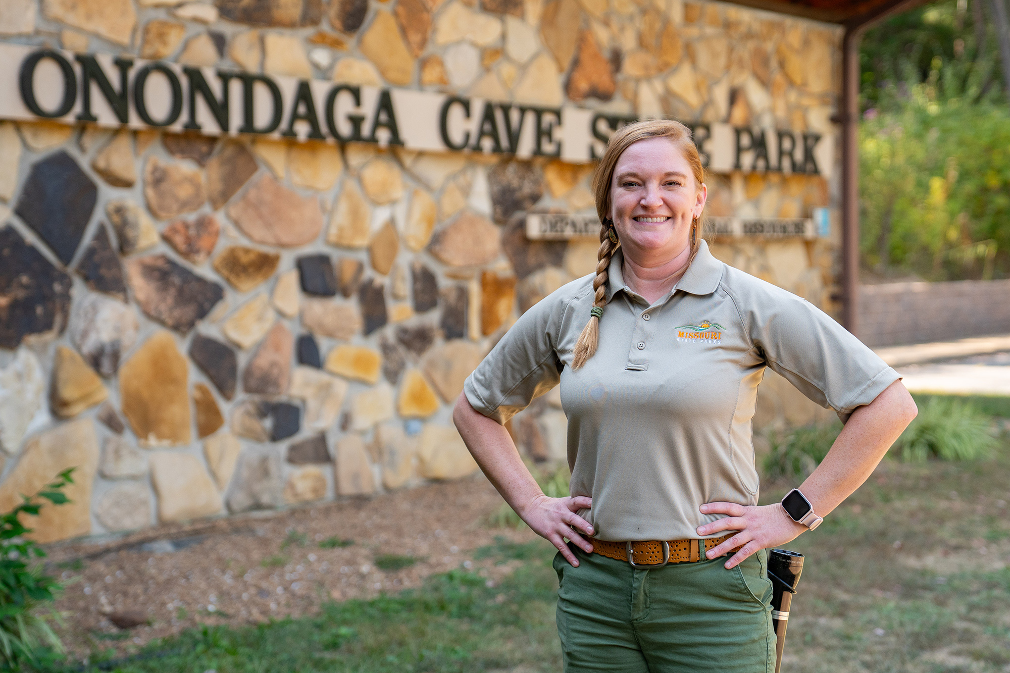 A woman in a state park shirt stands smiling at the camera with her hands on her hips. Behind her is the Onondaga Cave State Park sign.