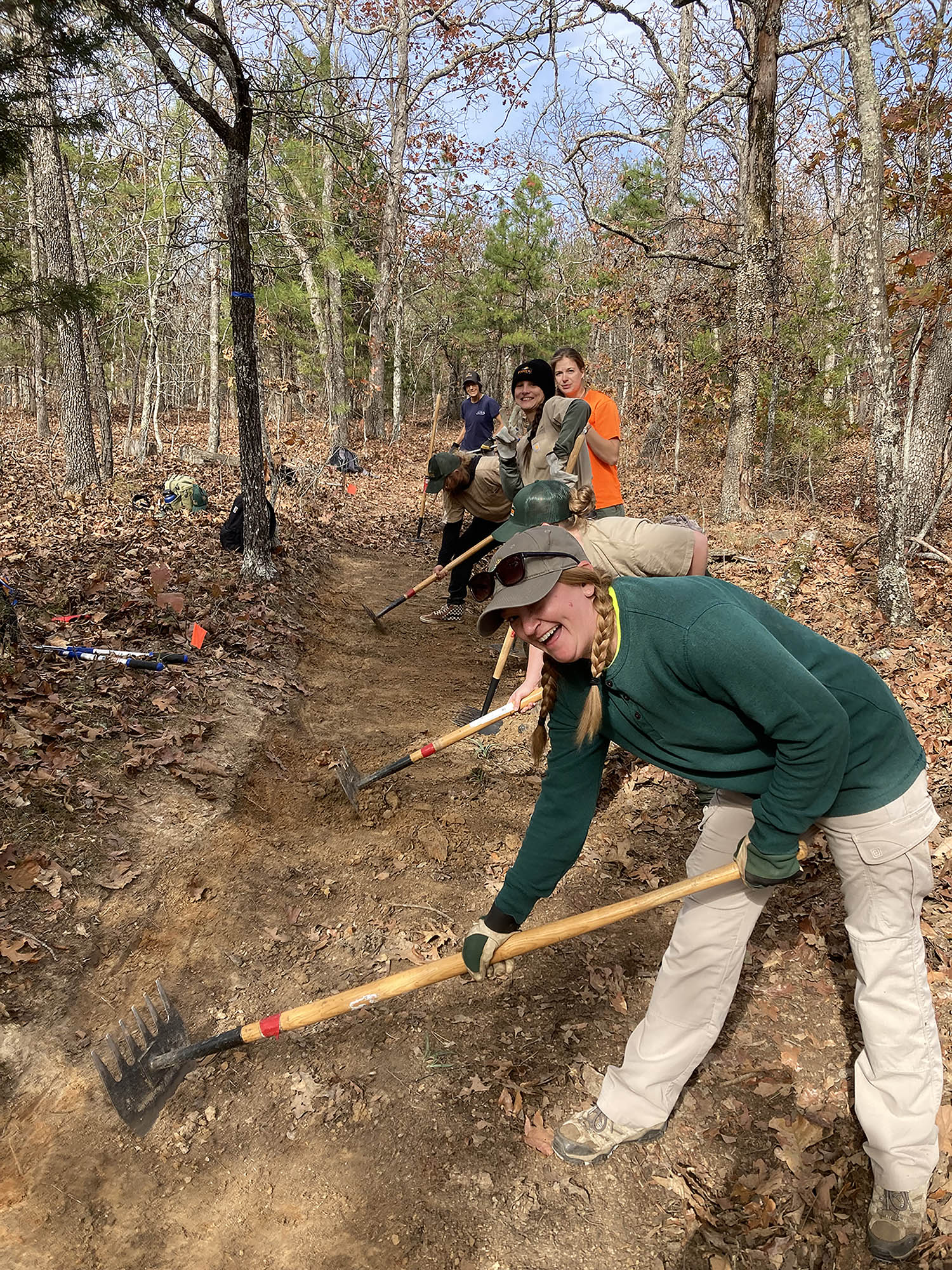 Six people in a row work with rakes to clear and level a dirt path through autumn trees and foliage.