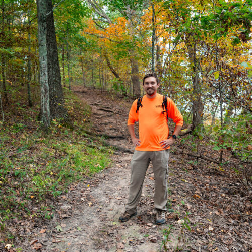 A man stands smiling at the camera with his hands on his hips, behind him a trail curves around the bend obscured by early autumn tress and undergrowth.