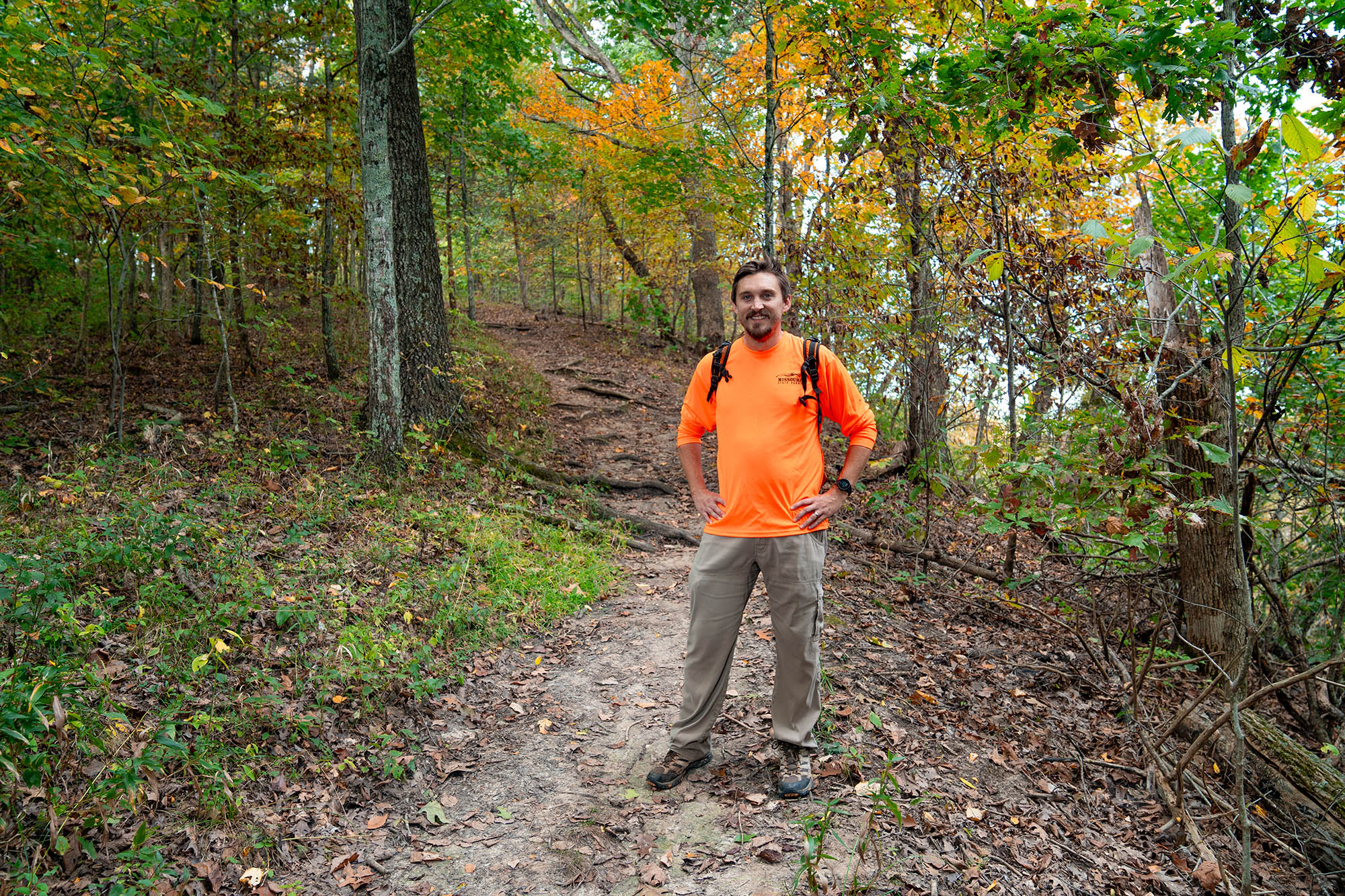A man stands smiling at the camera with his hands on his hips, behind him a trail curves around the bend obscured by early autumn tress and undergrowth.